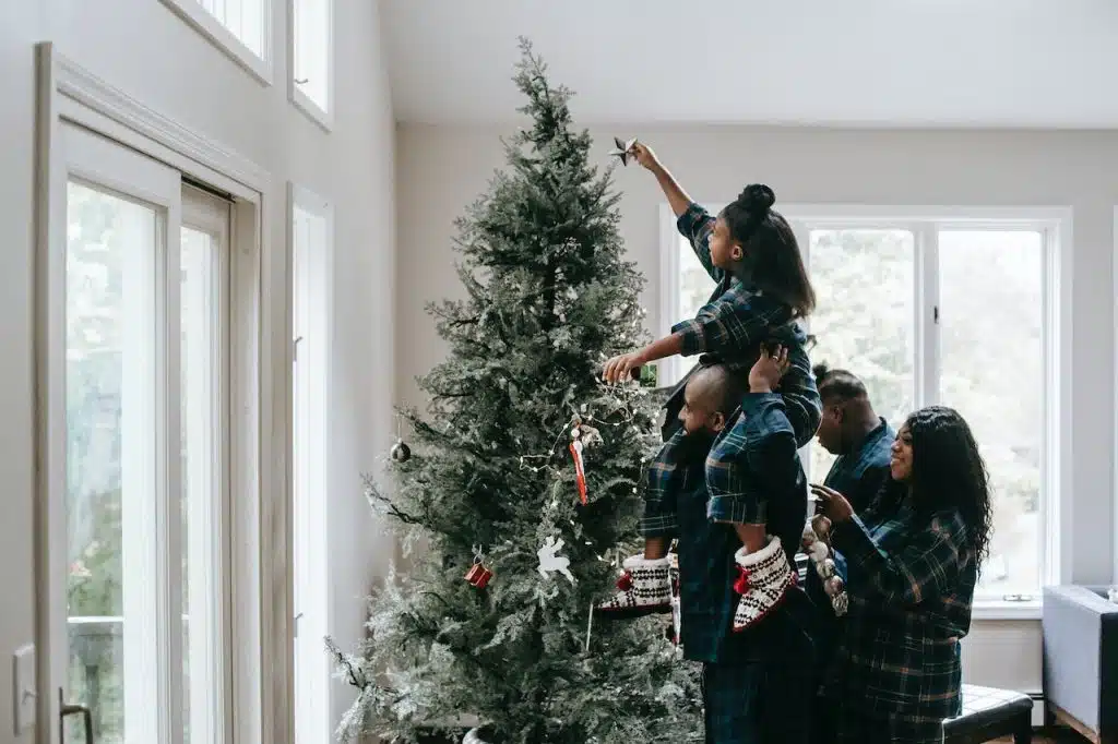 Un papa porte sa fille sur ses épaules pour qu'elle mette l'étoile au sommet d'un sapin de Noël. La maman se tient juste derrière eux.