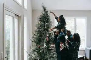 Un papa porte sa fille sur ses épaules pour qu'elle mette l'étoile au sommet d'un sapin de Noël. La maman se tient juste derrière eux.