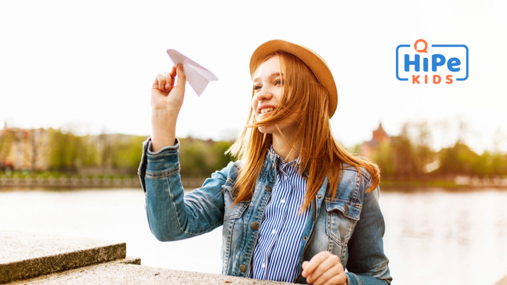 Une adolescente rousse avec un chapeau est assise à une table en extérieur et tient un avion en papier dans la main droite.
